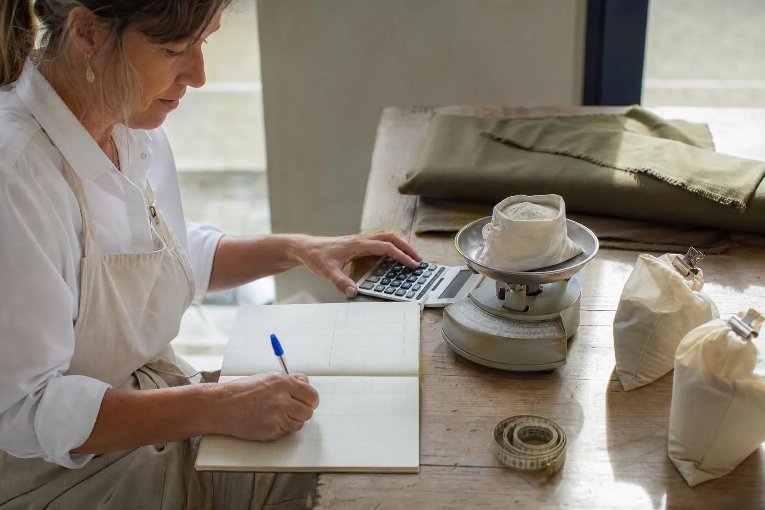 Femme calculant un devis à la main sur un carnet à colonnes, calculatrice et sachets de colorant pesés à côté